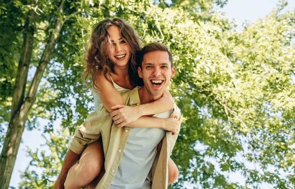 Pareja feliz divirtiéndose afuera en el parque. Novio dando paseo a cuestas en hombro a su bonita novia en unas vacaciones de verano en la naturaleza —  Fotos de Stock