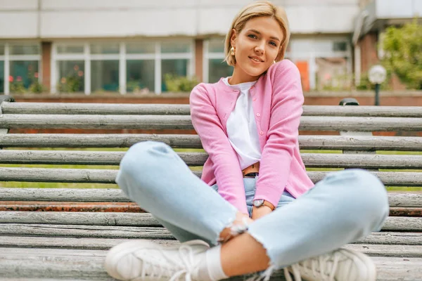 Mooie blonde jonge vrouw die breed glimlacht, een blauwe jeans draagt, een wit t-shirt en een roze jasje, glimlacht en zit op de bank in de straat. Gelukkig vrouwtje rustend buiten in de lente. — Stockfoto