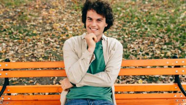 Positive young man with curly hair, smiling and looking to the camera, feeling joyful, sitting on the bench on the city street outdoors. Happy smart student male has happy expression.