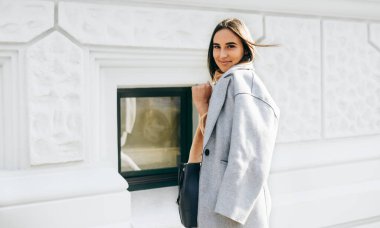 Image of beautiful young brunette woman wearing casual outfit, smiling and looking directly to the camera. Pretty female student walking in the city street. 
