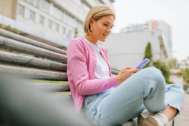 Side view image of pretty blonde young woman wearing blue jeans, white t-shirt and pink jacket, smiling and sending messages on smart phone sitting in the bench in the city street in spring time.