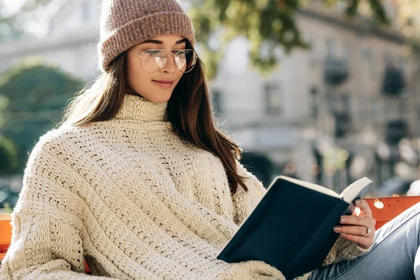 Image of young woman wearing sweater, hat and transparent eyeglasses, sitting on the bench in the city street and reading the book while waiting her friends. Young female student learning outside. — Stock Photo, Image
