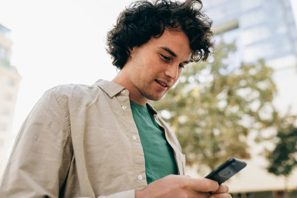 Caucasian smart young man standing outdoors, typing messages on mobile phone. Young handsome male with curly hair browsing online on his smart phone in the city street. — Stock Photo, Image