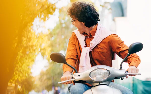 Beeld van vrolijke jonge zakenman met krullend haar rijdend op zijn scooter in de straat van de stad, terugkijkend, gekleed in casual outfit en transparante bril, heeft vrolijke uitdrukking. — Stockfoto