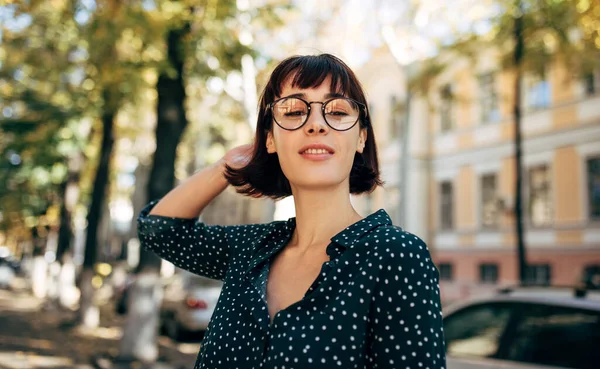 Utendørsbilde av en vakker ung kvinne i grønn skjorte med hvite prikker og gjennomsiktige briller, smilende og i kameraet. Ung kvinnelig student som poserer i bygaten . – stockfoto