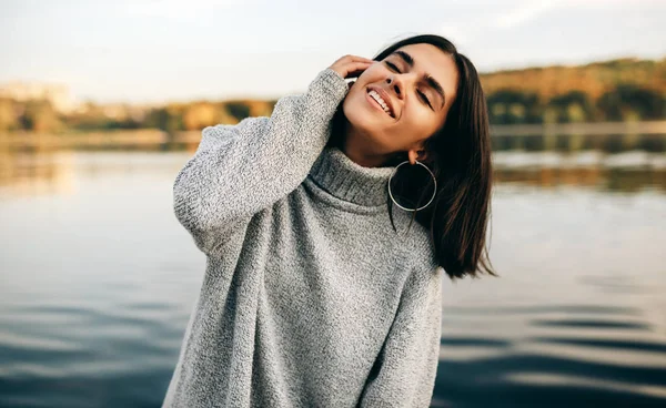 Зображення щасливої молодої брюнетки з закритими очима, одягнена в сірий светр, позує на фоні природи. Красива дівчина позує біля озера в парку . — стокове фото
