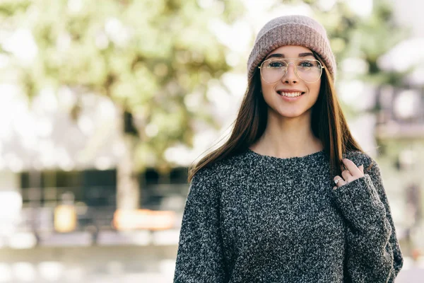Портрет молодої жінки, яка посміхається, одягнена в прозорі окуляри, сірий светр і рожевий капелюх. На відкритому повітрі зображення красивої студентки дивиться на камеру і розслабляється на міській вулиці . — стокове фото