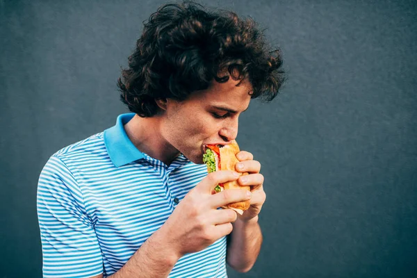 Egy jóképű fiatalember egészséges hamburgert eszik. Éhes ember egy gyorsétteremben hamburgert eszik a szabadban. Férfi göndör hajjal, utcai kajával és hamburgerrel. — Stock Fotó