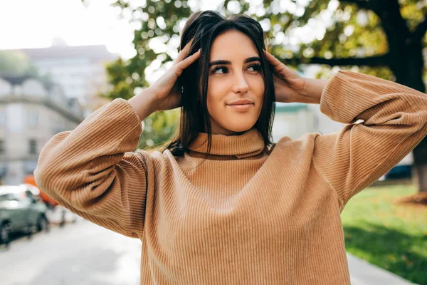 Retrato ao ar livre de sorrir bela jovem morena vestindo camisola bege, sorrindo e olhando para um lado. Estudante bonita posando na rua da cidade . — Fotografia de Stock