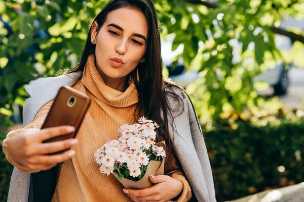Красива молода жінка говорить про власний портрет з квітами на мобільному телефоні, відкритому проти цегляної стіни. Гарненька брюнетка жінка стоїть на вулиці міста робить селфі . — стокове фото