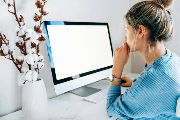 クリエイティブオフィスでデスクトップコンピュータを使用してフリーランスのビジネス女性の側面図。若いブロンドの女性の写真はあなたのテキストメッセージやコンテンツのための空白の画面を持つコンピュータ上で自宅で作業中に屋内に座る — ストック写真