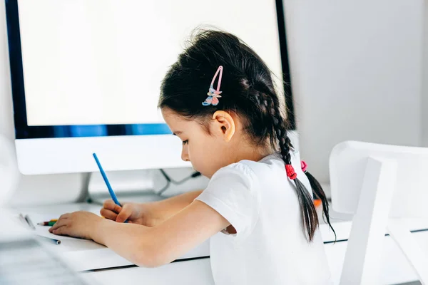 Evde bilgisayarın yanında kalemlerle resim yapan sevimli kız çocuğu. Odada bir masada oturan yaratıcı çocuk resim yapmayı öğreniyor. Küçük kız evde ödev yapıyor.. — Stok fotoğraf