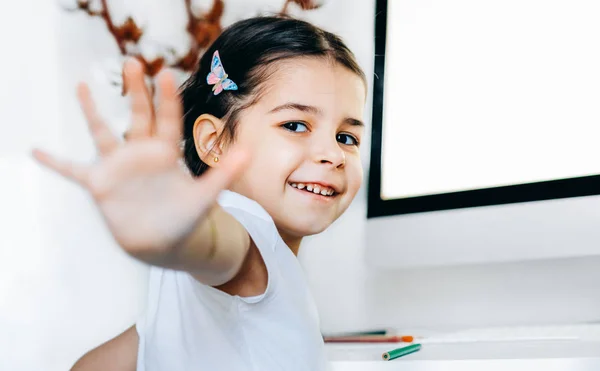 Glücklich schönes Kind Mädchen macht Geste hoch fünf sitzt neben dem Computer zu Hause. entzückendes Kind, das an einem Tisch im Zimmer sitzt und lernt. Kleinkind macht Hausaufgaben zu Hause. — Stockfoto