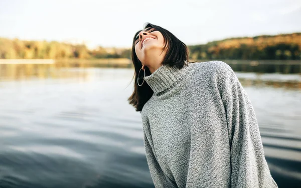 Imagem de jovem morena feliz com os olhos fechados, vestindo camisola cinza, posando no fundo da natureza. Menina bonita posando contra o lago no parque . — Fotografia de Stock