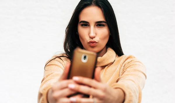 Portrait de magnifique jeune femme parlant autoportrait et soufflant un baiser sur un téléphone portable contre un mur blanc. Jolie brune debout dans la rue de la ville faisant selfie . — Photo