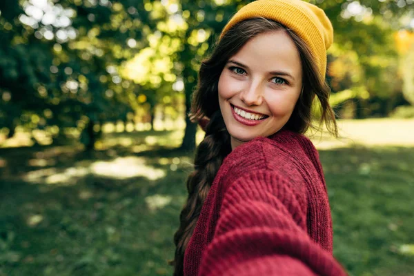 Linda jovem loira sorrindo amplamente com dentes brancos saudáveis, vestindo uma camisola vermelha e um chapéu amarelo, tirando um auto-retrato no fundo da natureza. Feliz fêmea fazendo selfie no smartphone . — Fotografia de Stock