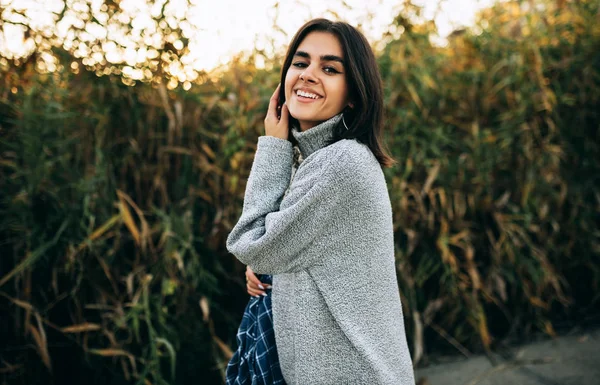 Mulher feliz posando em fundo verde, olhando para a câmera tem expressão alegre. Mulher morena jovem bonita, vestindo camisola cinza, posando no fundo da natureza . — Fotografia de Stock