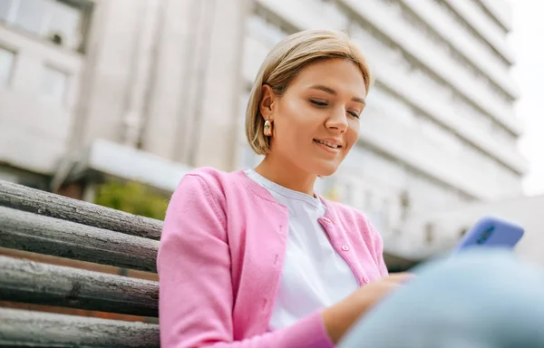 Imagem de jovem loira inteligente vestindo camiseta branca e jaqueta rosa, sorrindo e enviando mensagens em um smartphone. Bela navegação feminina na web na rua da cidade na primavera . — Fotografia de Stock