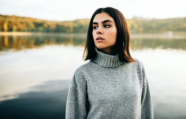 Молода жінка в сірому светрі, позує на фоні природи. Красива дівчина дивиться з одного боку, позує біля озера в парку . — стокове фото