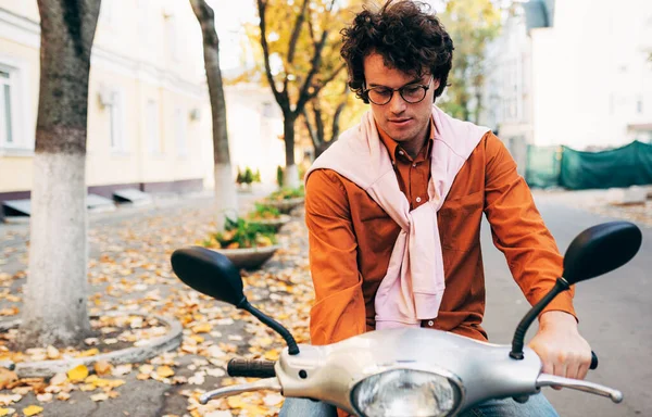 Mobiletiyle gezen zeki bir genç adam, günlük kıyafetler ve şeffaf gözlük takıyor, kıvırcık saçlı, ciddi bir ifadeye sahip..