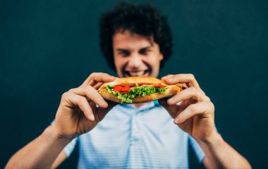 Çizburger yiyen genç bir adam. Fast-food restoranında açık havada hamburger yiyen mutlu bir adam. Kıvırcık saçlı bir adam sokak yemeği yiyor ve hamburger yiyor..
