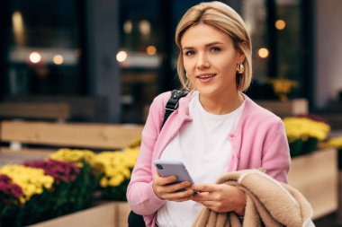Beyaz tişört ve pembe ceket giyen çekici genç bir kadın. İnternetten alışveriş yaparken gülümsüyor ve sokakta yürüyor. Güzel sarışın kız öğrenci cep telefonundan mesaj yolluyor..