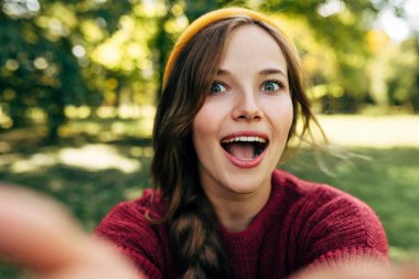 Geniş bir gülümsemeyle gülümseyen mutlu sarışın kadının yakın plan portresi, parktaki doğa arka planında poz veren şaşırtıcı bir ifadeye sahip. Selfie çeken güzel bir kadın. İnsanlar, seyahat, yaşam tarzı