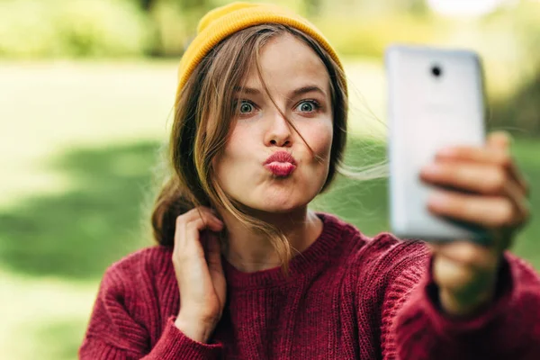 Güzel sarışın bir kadının açık hava portresi cep telefonuyla arkadaşına öpücük yolluyor, kırmızı kazak giyiyor, sarı şapkalı, parkta doğa arka planında poz veriyor. Mutlu kadın selfie çekiyor..
