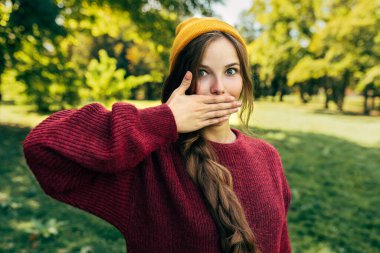 Genç sarışın bir kadın, süveter ve sarı bir şapka giyerek, dudaklarını oldukça uzun süre kapatıyor. Parktaki doğal arka planda poz veriyor. Mutlu kadın dışarıda poz veriyor, ağzını kapatıyor, sessiz jest yapıyor..