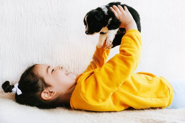 Evde küçük bir köpekle oynayan gülümseyen mutlu bir çocuğun yan görüntüsü. Güzel küçük kız köpek yavrusunu umursuyor. Sevimli bir çocuk güneşli bir günde halıda evcil hayvanıyla oynuyor..