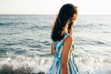 Güneşin battığı arka planda kumsal ve deniz boyunca yürüyen esmer bir kadının görüntüsü. Elbise giyen, okyanusta yürüyen güzel bir kadın..