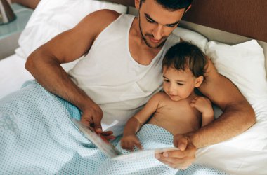 Baba ve kızı yatakta uzanıyor ve evde birlikte kitap okuyorlar. Evdeki sevimli bebeğe peri masalları anlatan bir baba tasviri. Şirin küçük kız ve yakışıklı babası kitap okuyorlar..