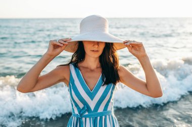Sahilde yürüyen güzel bir kadının yakın plan portresi tatil sırasında sıcak güneşin tadını çıkarıyor. Çizgili elbise giyen ve gözlerini okyanus arka planında büyük beyaz şapkayla kapatan güzel bir kadın manken..