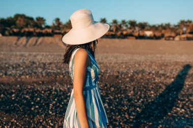 Sahilde gün batımında arka planda yürüyen esmer, beyaz şapkalı güzel bir kadının yan görüntüsü. Tatil sırasında dışarıda poz veren çizgili elbiseli bir kadın. Seyahat kavramı. 