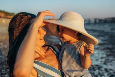 Mutlu gülümseyen genç anne ve kızının gün batımında eğlenirken yan görüntüsü. Güzel bir dişi ve bebek gün batımında okyanus sahilinde birlikte oynuyorlar. Annelik aşkı bakımı.