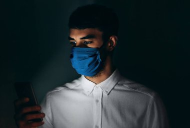 Siyah arka planda virüs salgını sırasında akıllı telefon kullanarak tıbbi mavi maske takan bir adamın görüntüsü. Cep telefonu kullanan, tek kullanımlık maske takan beyaz bir erkek. Coronavirus, sağlık hizmetleri..