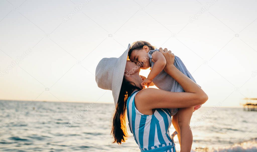 Image of happy young mother playing with her daughter at the sea sunset. Beautiful female kissing baby walking outside at the ocean beach on sunset. Motherhood love care.