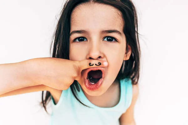 Amazed little girl makes grimace with wide open mouth, with mustache drawing on her index finger put it under the nose. Funny child having fun and playing inside. Human expressions, children concept