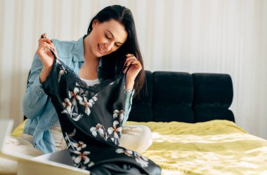 Yatağa oturmuş gülümseyen ve paketini yeni bir elbiseyle açan genç bir kadın görüntüsü. Mutlu kadın evde doğum günü hediyesi alıyor. Çevrimiçi internet ticaret dağıtım kavramı