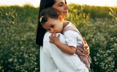 Açık havada gülümseyen anne ve tatlı küçük kızın gün batımında sahada birbirlerine sarıldıkları görüntüler. Anne ve kızı doğa arka planında bir çiçek tarlasında yürüyor..