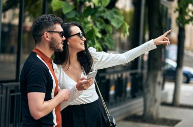 Şehir sokaklarında akıllı telefondan bir şey doğrultmuş genç bir kadın ve bir adamın açık hava görüntüsü. Genç çift harita uygulaması için cep telefonunu kullanarak şehir caddesinde yürüyor..