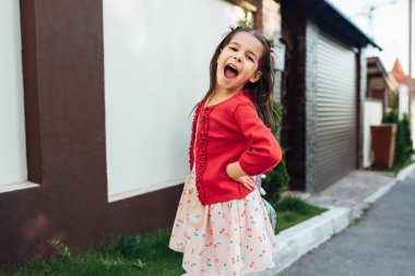 Okuldan sonra eve gelirken evinin önünde duran sevimli küçük bir kızın gülüşü. Dışarıda oynayan gülümseyen çocuğun neşeli bir ifadesi var. Mutlu çocuk gülümseyerek ve dışarıda poz vererek