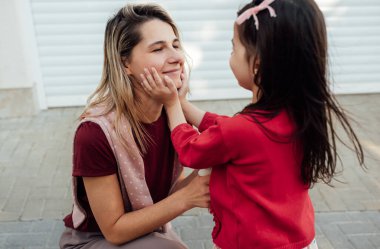 Küçük sevimli bir kızın açık hava görüntüsü. Gülümseyen annesinin yüzüne şefkatle sarılıyor. Anaokulundan sonra kızı annesine sarılıyor. Mutlu bir kadın evinin önünde çocuğunu kucaklıyor..