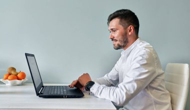 Bilgisayarda arkadaşlarıyla sohbet etmek için çalışan, internette gezinen, gülümseyen sakallı bir adamın yan görüntüsü. Mutfakta masada oturan yakışıklı erkek, bilgisayarda web sitesinde çalışıyor..