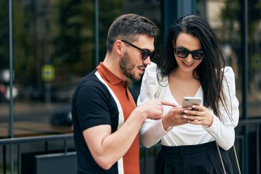Şehir sokaklarında akıllı telefon okuyarak duran genç kadın ve adamın yan görüntüsü. Genç çift harita uygulaması için cep telefonunu kullanarak şehir caddesinde yürüyor..