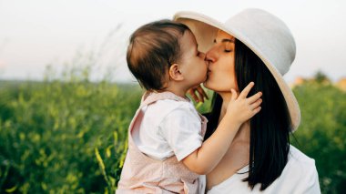 Güzel anne ve tatlı küçük kızın yakın plan portresi. Gün batımında, açık havada birbirlerine sarılıyorlar. Anne ve kızı çiçeklerle dolu bir tarlada oturuyor..