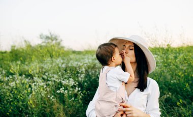 Güzel bir anne ve küçük tatlı bir kızın gün batımında, sahada birbirlerine sarıldıkları yatay bir görüntü. Anne ve kızı çiçeklerle dolu bir tarlada oturuyor..