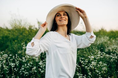 Açık havada, beyaz şapkalı, sahada yürüyen muhteşem bir kadının portresi. Mutlu kadın gülümsüyor ve yeşil çimlerin üzerinde bir yaz gününün tadını çıkarıyor..