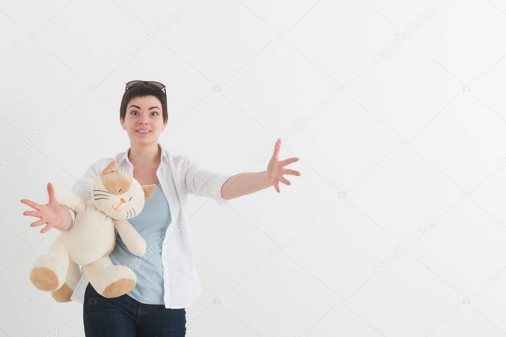 Portrait of a young girl in white shirt on the light background. Looking at the camera, smiling and extending hands forward to the spectator. Happy woman with plush toy cat