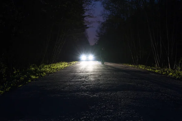 Erkek siluet gece ormanda bir karanlık dağ yolda kenarında. Adam yolda araba farlar karşı ayakta. Araba yol üzerinde. Gizem kavramı. Yumuşak odak, filtre.